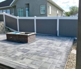 A grey stone paver patio featuring a rectangular brick fire pit in front of a white and grey vinyl fence.