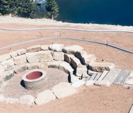 A circular stone fire pit and seating area overlooking a lake.