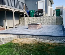 A newly installed gray paver patio with a central brick fire pit, bordered by a tiered retaining wall.