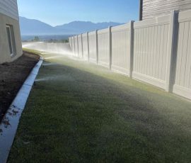Lawn sprinklers watering the grass alongside a white vinyl fence.
