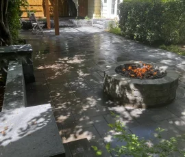 A stone patio with a fire pit, a built-in stone bench, and a wooden pergola near a house.