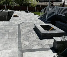 A backyard patio with gray stone pavers, a built-in fire pit, and stairs leading up to a house deck.