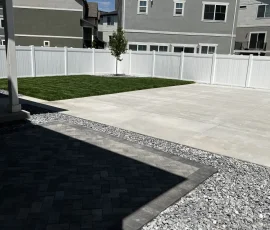 Backyard featuring a paved patio, a gravel border, a section of green lawn, and a white fence in front of modern houses.