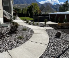 A concrete walkway curves through a backyard featuring gravel landscaping, a hot tub, and mountains in the distance.