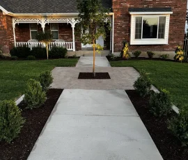 A brick house with a front porch, a sidewalk leading to the entrance, and landscaped garden beds with small trees.