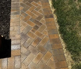 A herringbone-patterned brick walkway bordered by grass and mulch.