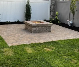 A square stone fire pit on a brick patio in a fenced backyard with a mountain view.