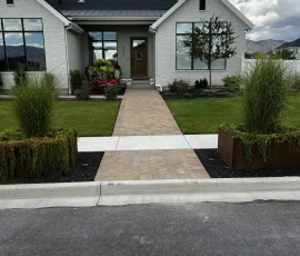 White brick house with a paved walkway, a front door, and two large metal planters with tall grasses.