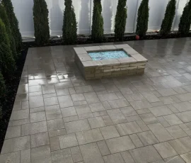 A square stone fire pit filled with blue glass beads on a paved patio bordered by tall evergreen trees and a white fence.