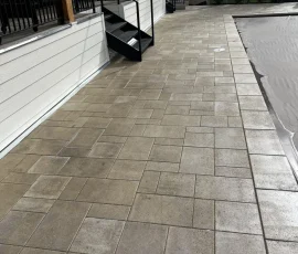 Paver patio beside a swimming pool with a black metal railing and stairs leading to a house deck.