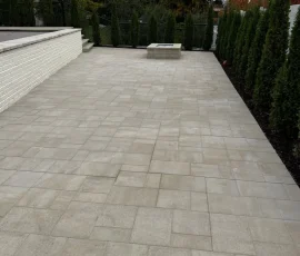 A stone paver patio featuring a low retaining wall and a fire pit, bordered by a line of tall evergreen trees.