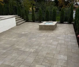 Backyard patio with stone pavers, a square fire pit, and steps leading to a raised platform.