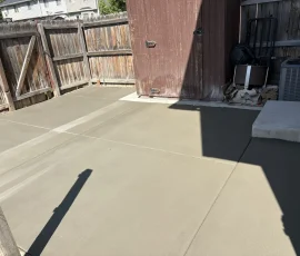 A newly poured concrete patio in a backyard next to a wooden fence and storage shed.