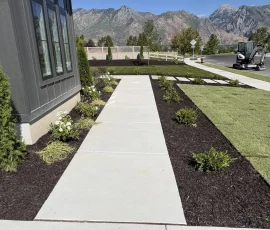 A concrete walkway leads through a landscaped yard with mulch beds and small shrubs, with mountains in the background.
