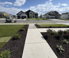 A concrete walkway leads through a landscaped yard toward modern suburban houses and a construction vehicle.