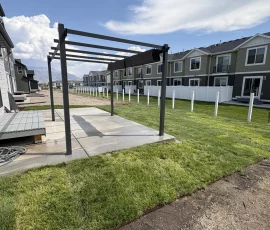A backyard with a concrete patio, a metal pergola, a wooden deck, and a newly laid lawn bordered by a white fence.