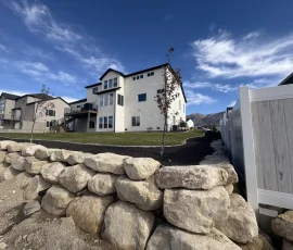 A modern two-story house with a white exterior, lawn, and a large boulder retaining wall in the foreground.