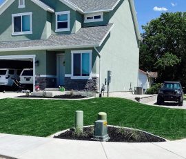 A two-story green house with a two-car garage, a green lawn, and two SUVs parked in the driveway and on the street.