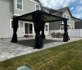Black metal gazebo with mesh curtains on a stone patio behind a house.