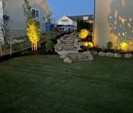 Stone steps leading up a sloped yard between two houses, illuminated by landscape lighting at dusk.