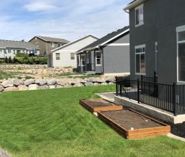 A gray house with a black railing, a green lawn, and two wooden raised garden beds.