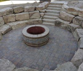 Circular fire pit in a stone-paved patio surrounded by a rock retaining wall and concrete steps.