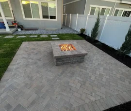 Backyard patio with a lit square stone fire pit and a herringbone paver pattern.