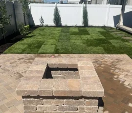 A square stone fire pit on a paver patio in a backyard with a newly installed sod lawn.