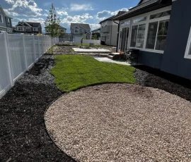 A backyard featuring a circular gravel patio, a patch of grass, and a white privacy fence.