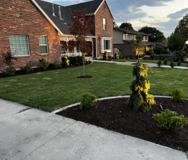 A brick house with a front lawn, landscaped garden beds, and young trees at dusk.