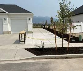A suburban driveway blocked off with yellow caution tape, featuring a workbench and newly planted trees.