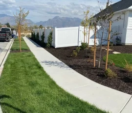 A white fence borders a sidewalk and freshly landscaped yard with young trees, with mountains in the background.