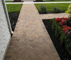 A herringbone brick walkway leading from a house porch to a street, flanked by landscaping and two white pumpkins.